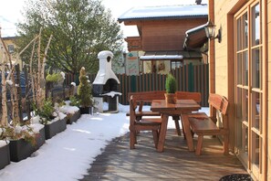 A wooden deck with a bench and a small table, surrounded by snow-covered plants and a wooden fence.