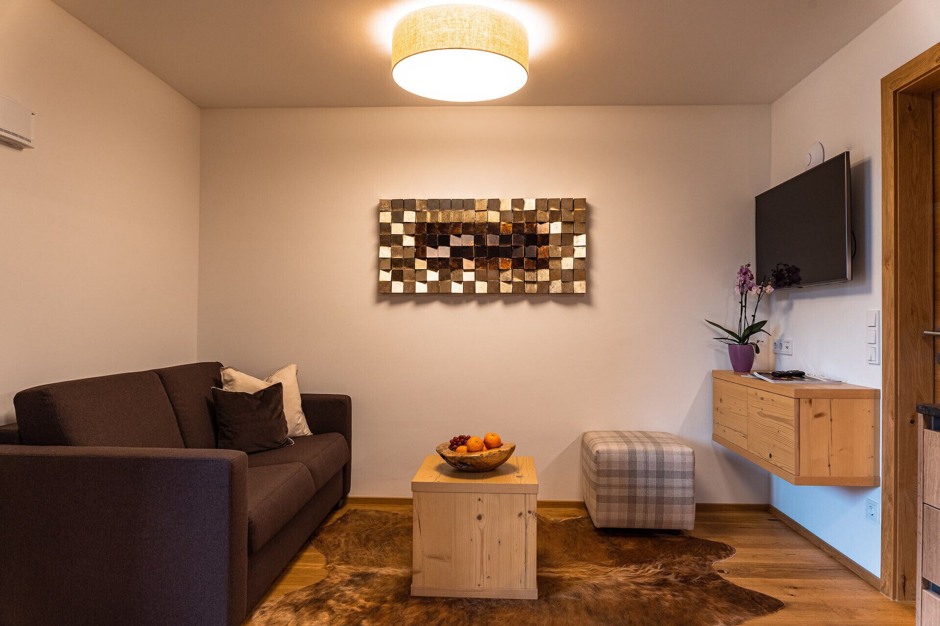 Una sala de estar con un sofá marrón, una mesita de madera con un cuenco de frutas, un reposapiés a cuadros, un adorno en la pared y un televisor montado.