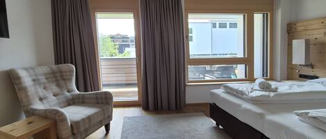 A bedroom with a bed, a chair, a small wooden side table, a window with curtains, and a view of buildings outside.