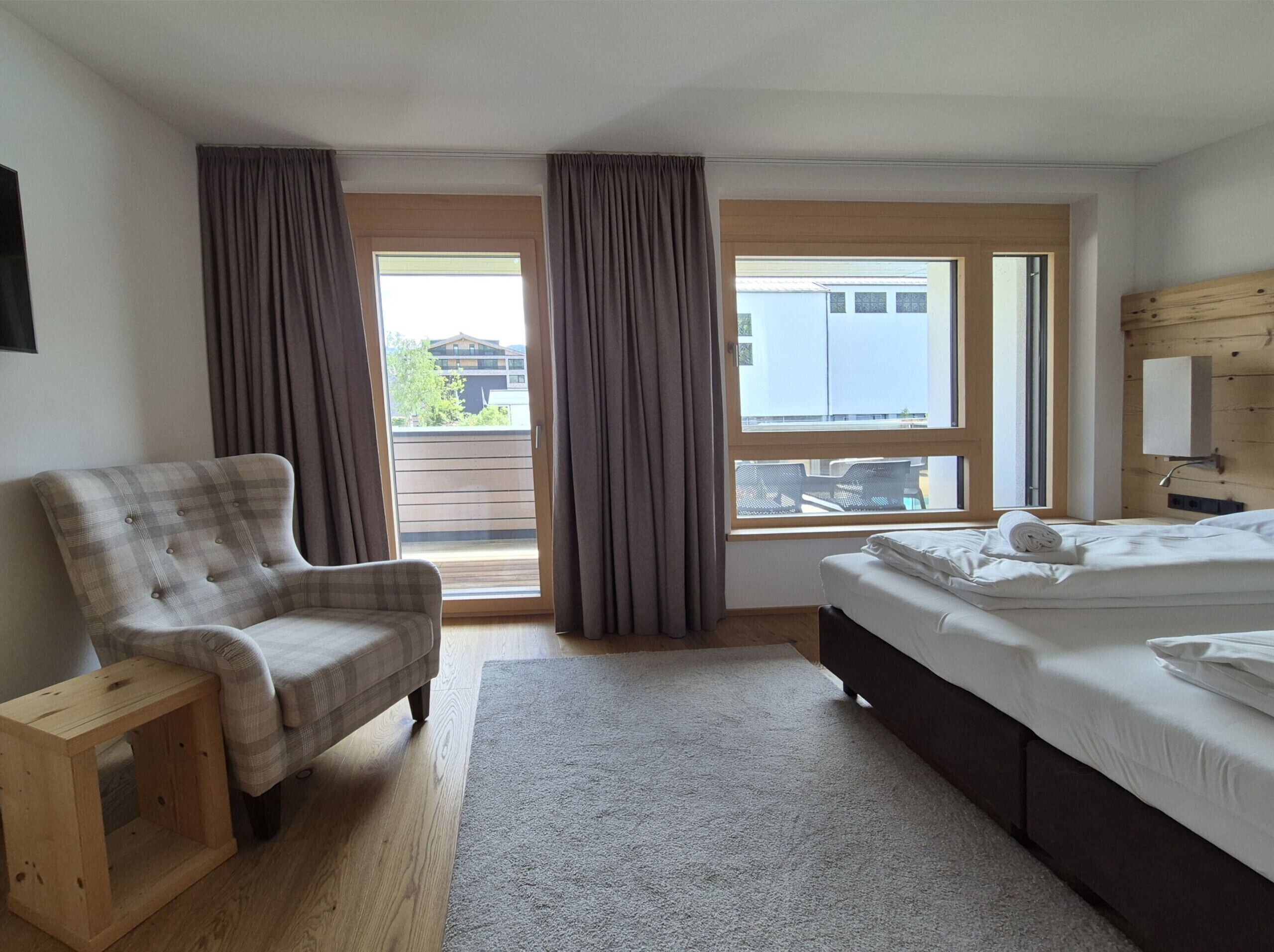 Un dormitorio con cama, una silla, una mesita de madera, un ventanal con cortinas y vistas a edificios.