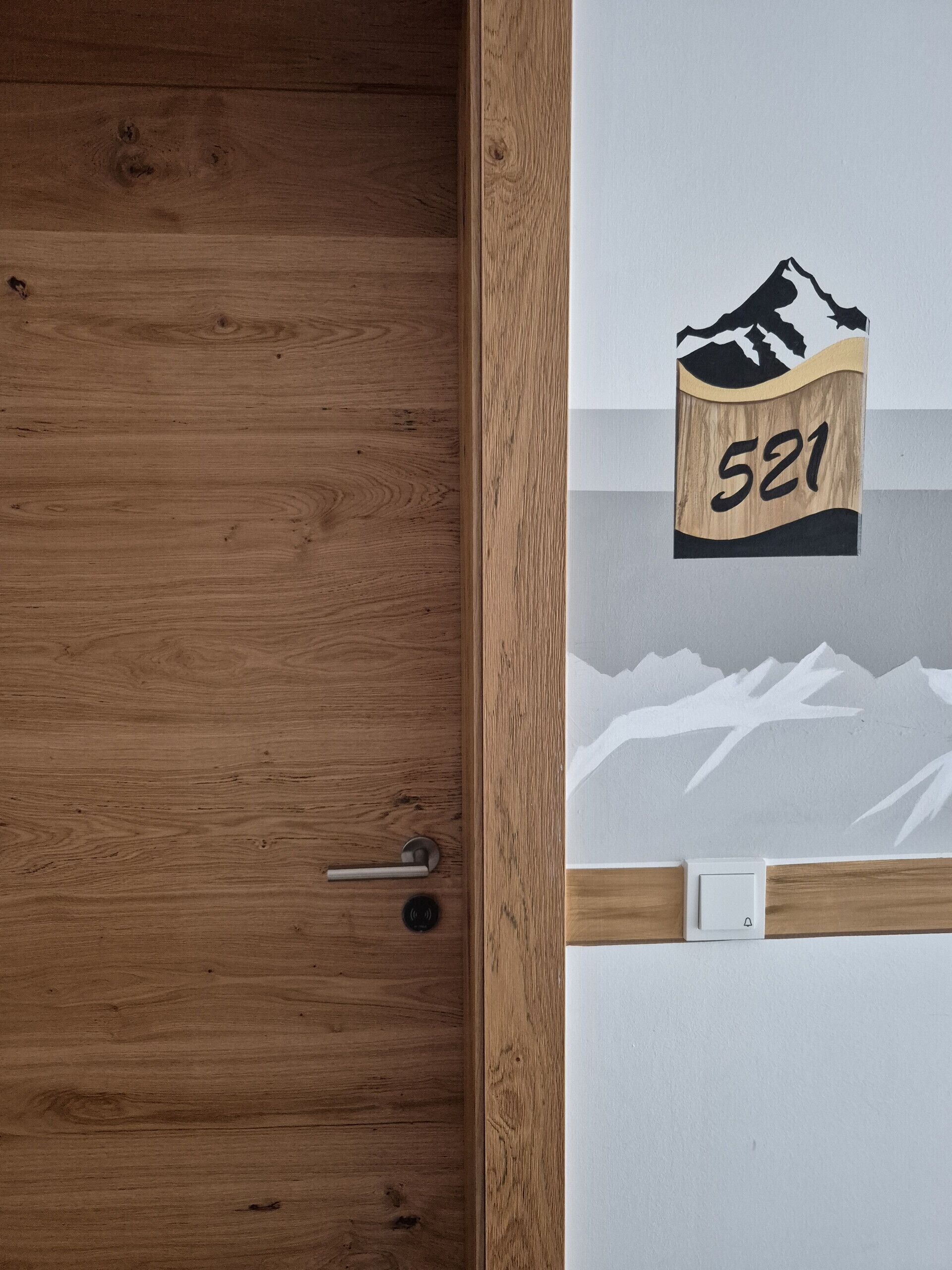 Una puerta de madera con manija metálica y una pared blanca con un adhesivo de un paisaje montañoso y el número '521'.
