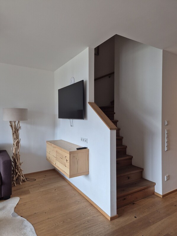 A modern living room with a flat-screen TV mounted on the wall, a wooden cabinet below it, a staircase leading to another floor, and a lamp on the floor. - SPA Apartments Deluxe Panorama 521 (Zell am See)