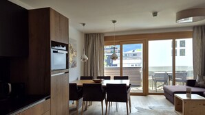A modern kitchen with wooden cabinets, a dining area with a table and chairs, a sofa, and a large sliding glass door leading to a balcony.