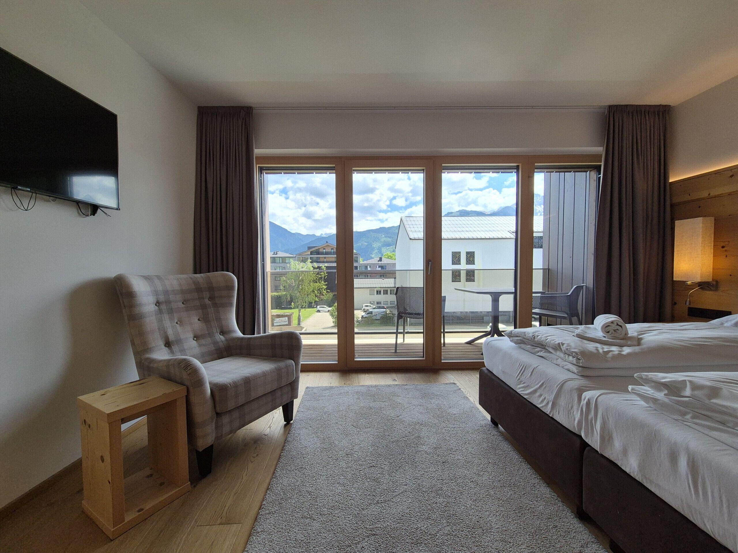 Habitación de hotel con cama, una silla, una mesita, televisor y balcón con vistas a edificios y montañas.