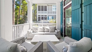 A sunlit porch with a white sofa and wicker chairs, a white table, and a view of trees and a house through the window. - WaterColor! Carriage House, Pool, LSV, Bikes! Next to Camp WaterColor Waterpark! (Santa Rosa Beach)