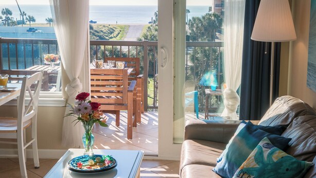 En balkon med udsigt over havet, et spisebord med stole, en sofa med puder og en lampe.