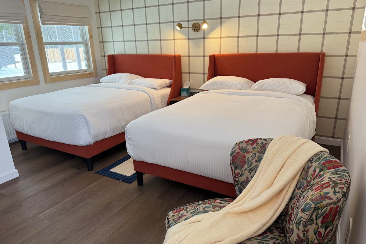 A hotel room with two beds, a sofa, a patterned armchair, and a window with a view of a fence.