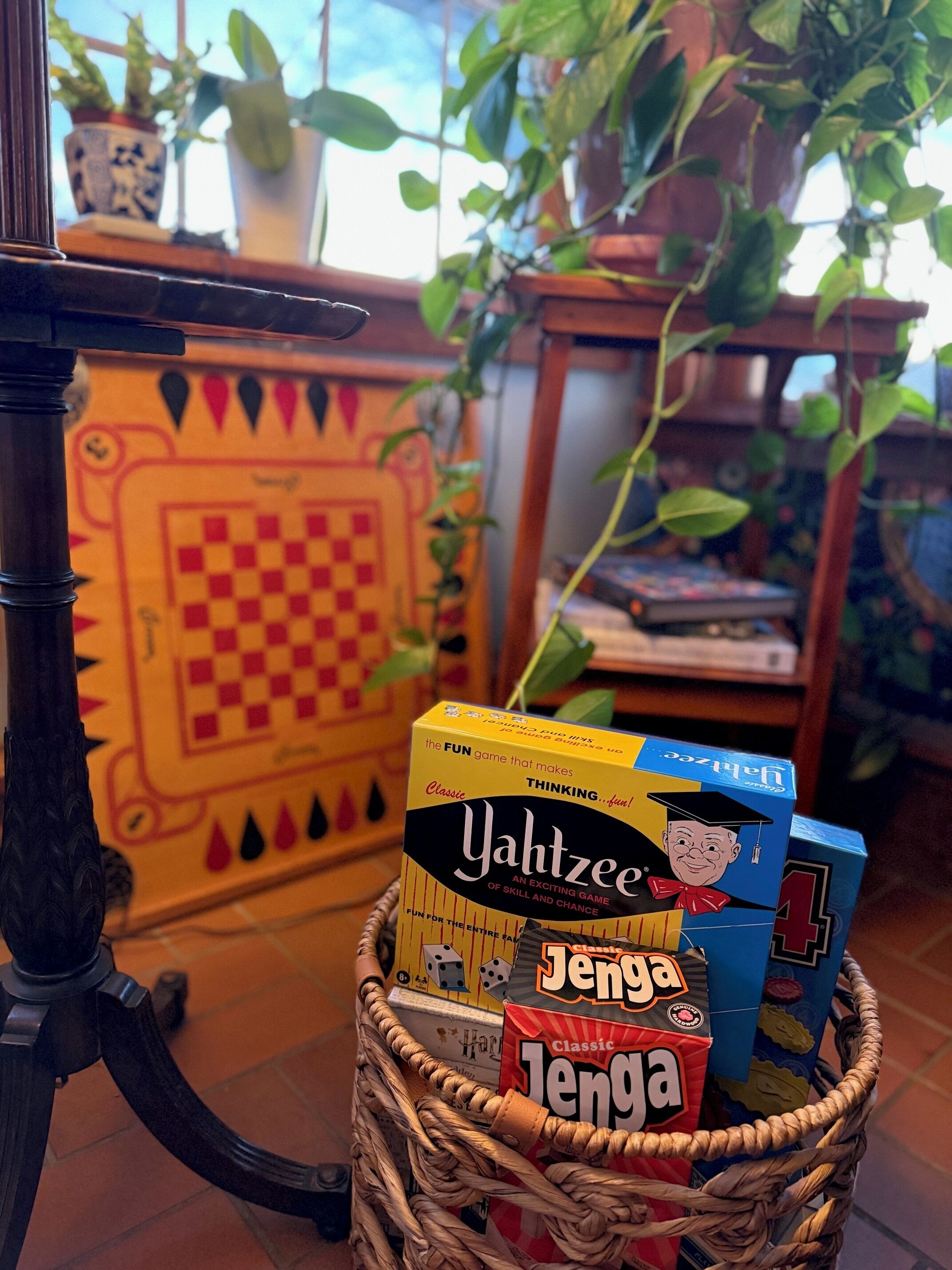 Uma cesta de vime com dois jogos de tabuleiro, 'Yachtzee' e 'Jenga', colocada sobre uma mesa com um tabuleiro de xadrez e plantas em vasos ao fundo.