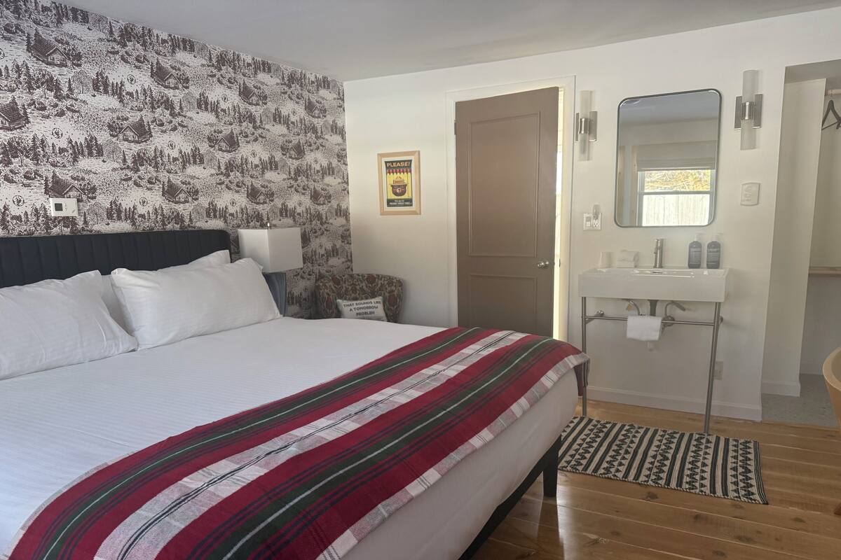 A bedroom with a large bed, a wooden floor, a bathroom with a sink and mirror, and a door.