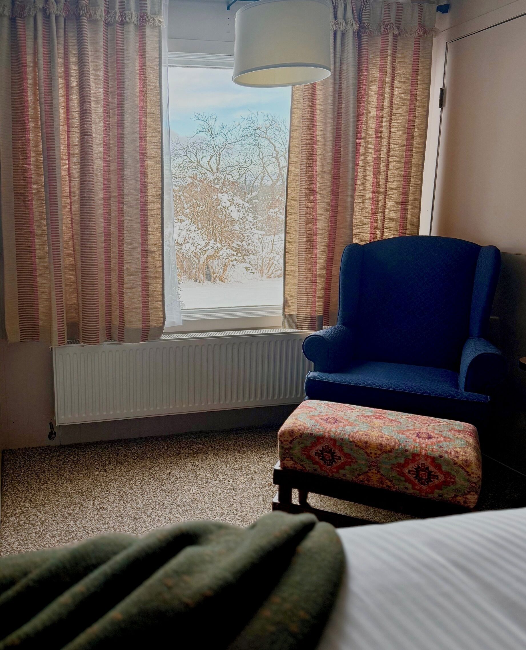 Una habitación con un sillón azul, un reposapiés estampado, un radiador y una ventana con cortinas.
