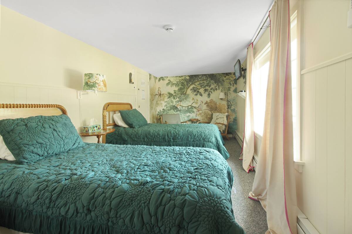 A room with two beds, each with a green quilt, a bedside table with a lamp, and a wall with a tree mural.