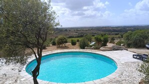 A circular pool with stone tiles, surrounded by rocks and trees, with a view of distant hills. - Li Scalitti Residence with swimming-pool (Badesi)