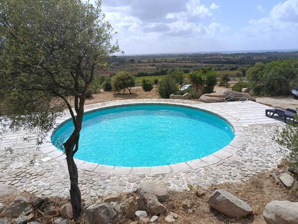 A circular pool with stone tiles, surrounded by rocks and trees, with a view of distant hills. - Li Scalitti Residence with swimming-pool (Badesi)
