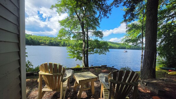 Blick auf einen See vom Erker aus, mit Adirondack-Stühlen und einem Tisch, mit Blick auf einen ruhigen See und bewaldete Hügel.