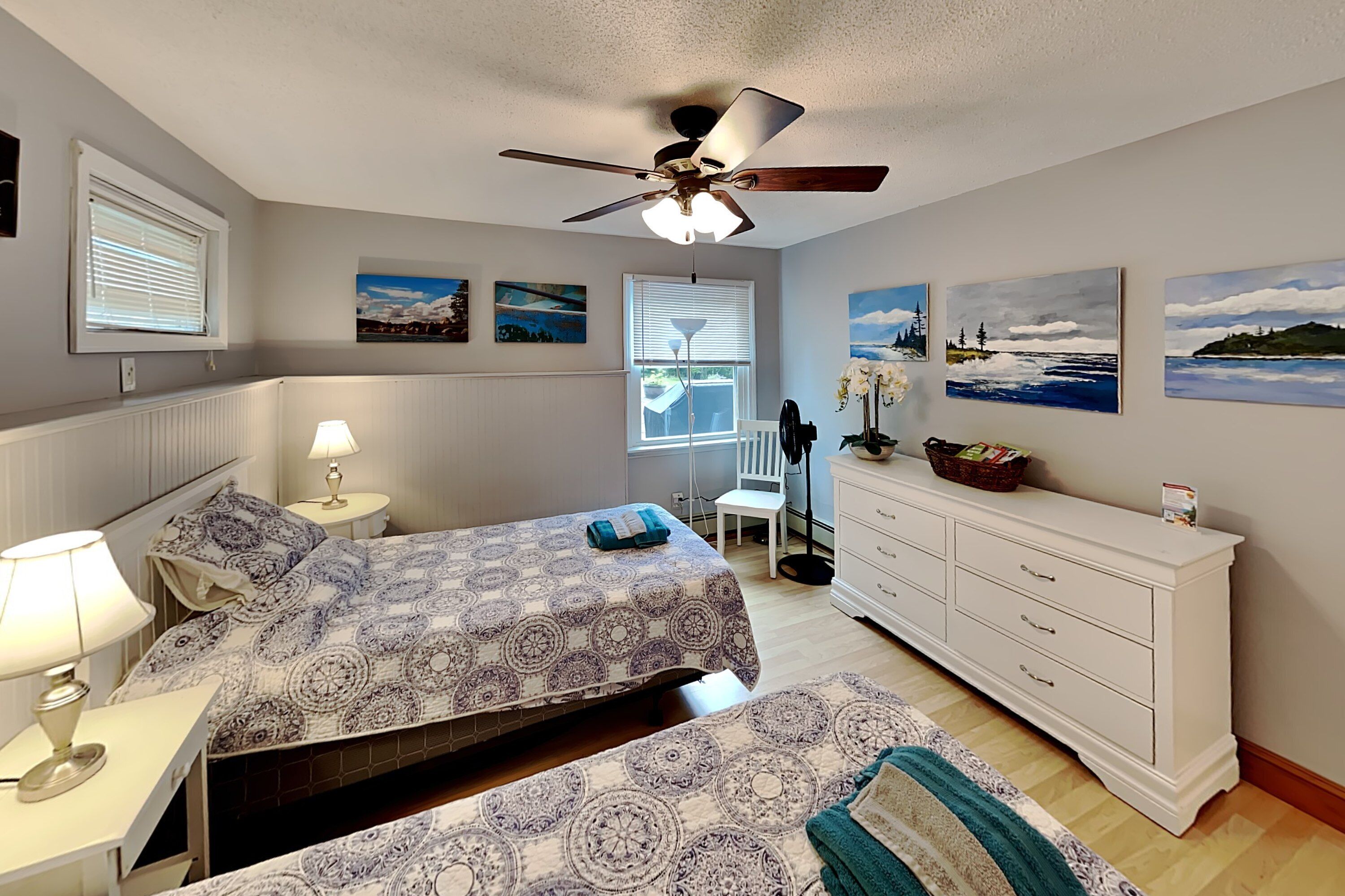 A bedroom with two beds, a ceiling fan, a dresser, and artwork on the walls.