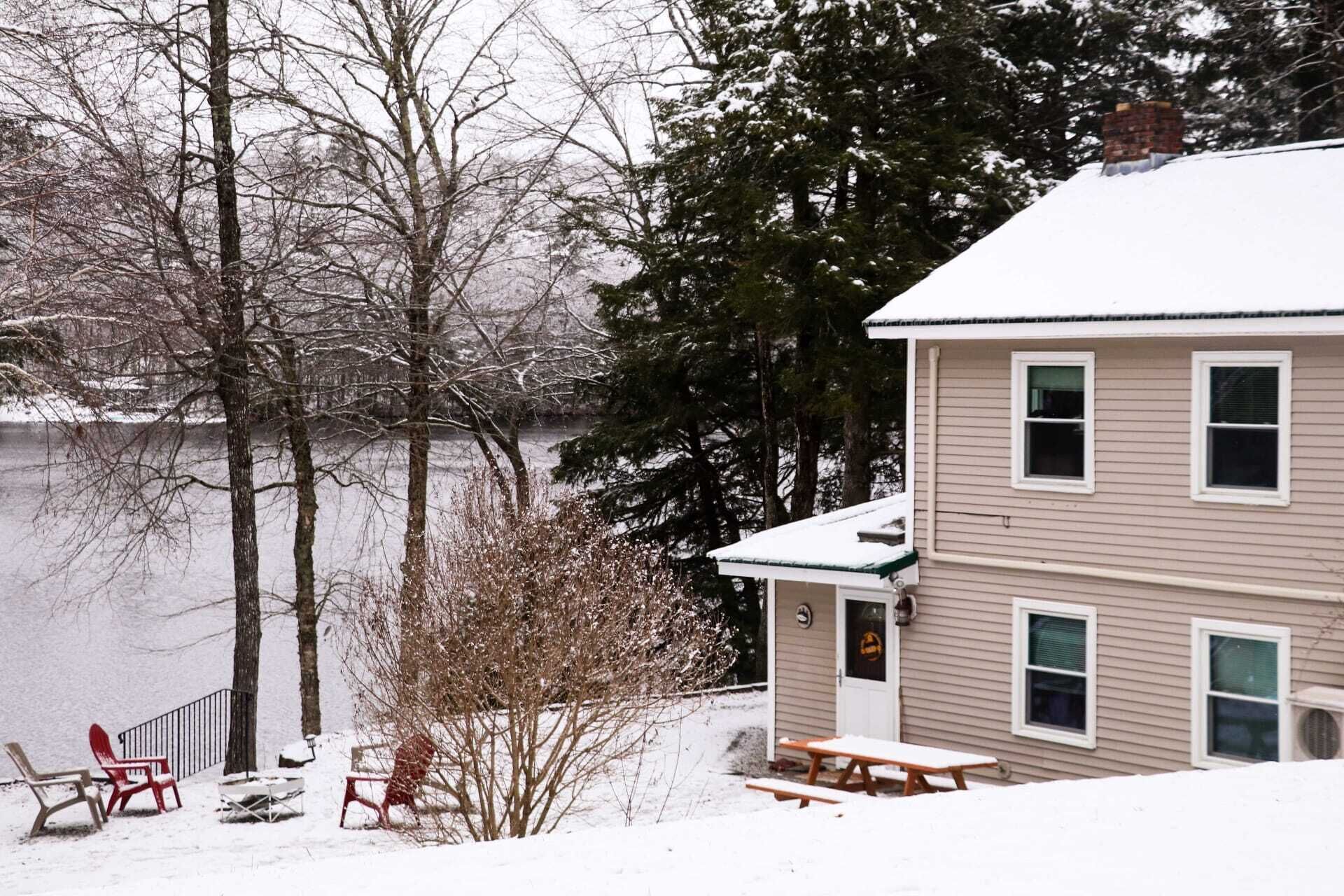 A two-story house with a snow-covered roof, a dock extending into a frozen lake, and a small deck with picnic tables.