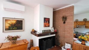 Una sala de estar acogedora con chimenea, un cuadro enmarcado y una zona de cocina rústica con armarios de madera y pared de ladrillo.