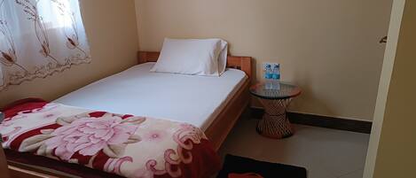 A neatly made bed with a floral bedspread, a bedside table with two water bottles, and a small round table with a woven base.
