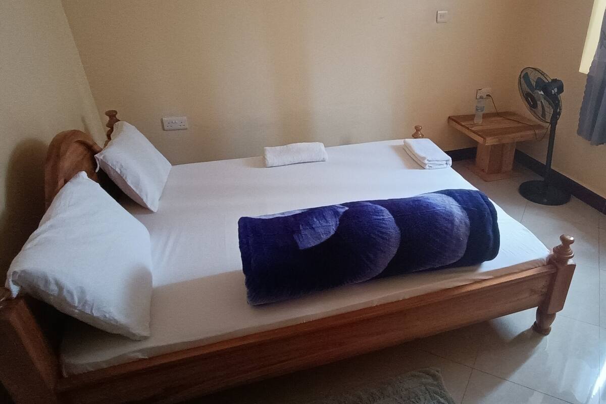 A neatly made bed with white linens and a blue blanket, a wooden nightstand with a fan, and a window with curtains.