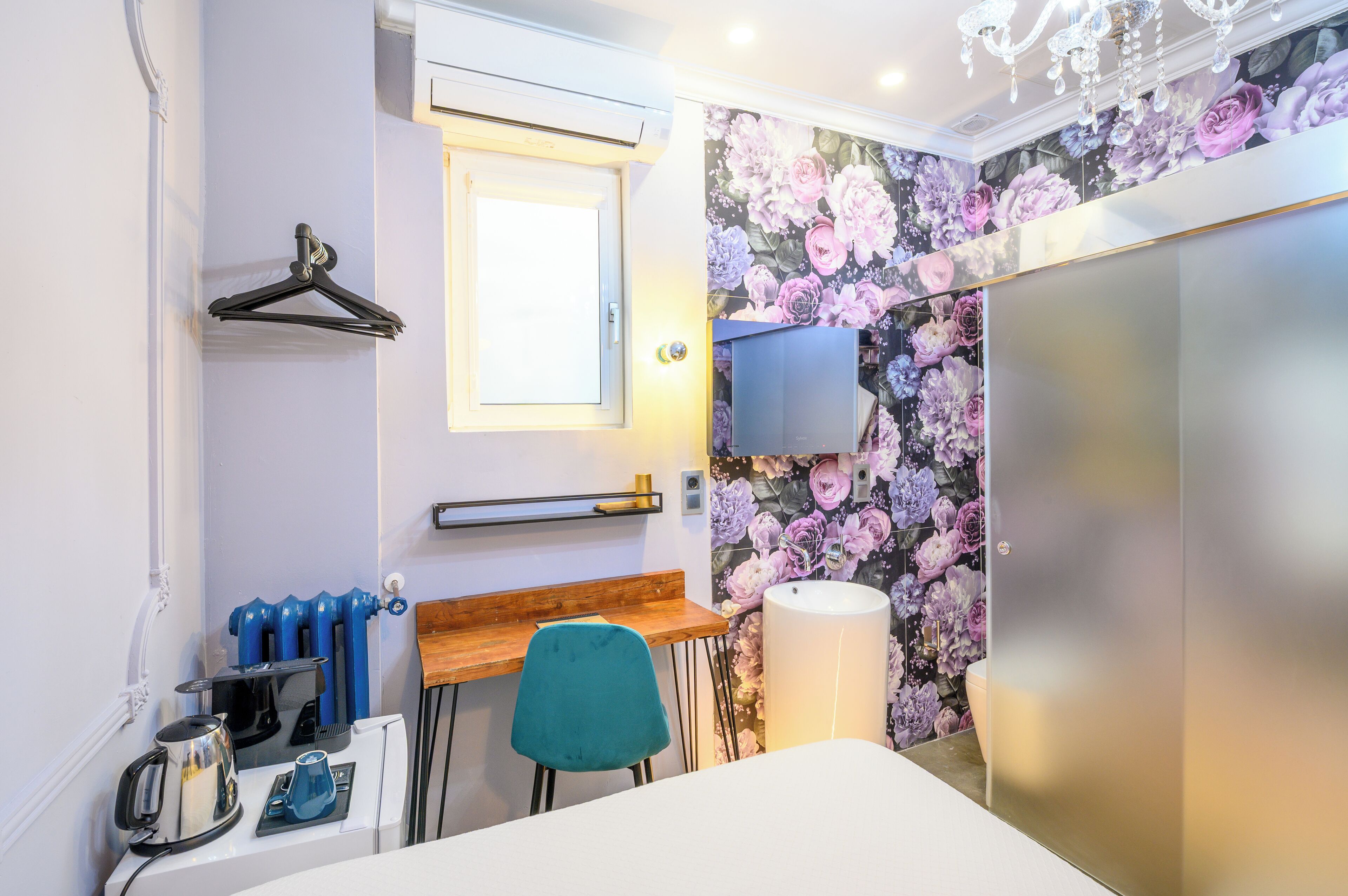 A compact hotel room with a wooden desk, a teal chair, a radiator, a kettle, a wall-mounted TV, and a floral wallpaper.