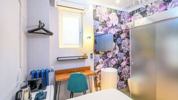 A compact hotel room with a wooden desk, a teal chair, a radiator, a kettle, a wall-mounted TV, and a floral wallpaper.