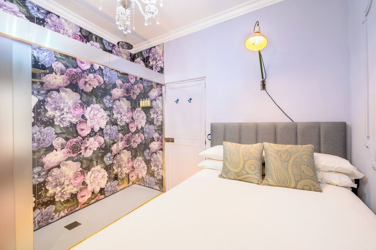 Ett sovrum med en blommig tapet, en säng med två kuddar, ett sängbord och en hängande lampa.