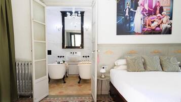 Een slaapkamer met een bed, een nachtkastje, een spiegel, een radiator, een badkamer met een bad en een kroonluchter.