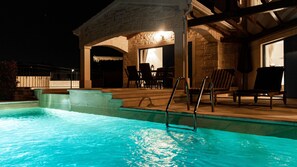 A swimming pool with steps leading into it, a covered patio area with chairs, and a stone building structure.