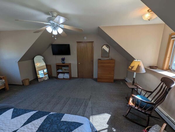 A bedroom with a sloped ceiling, a ceiling fan, a bed, a dresser, a chair, a lamp, a TV, and a door.