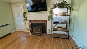 A living room with a fireplace, a television mounted above it, and a metal shelving unit with plants. - Brick House on the River at Riverside Point Resort (Park Rapids)