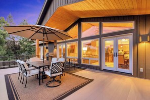 Een modern terras buiten met een eettafel en stoelen, een grote parasol en uitzicht op het interieur door grote glazen deuren.