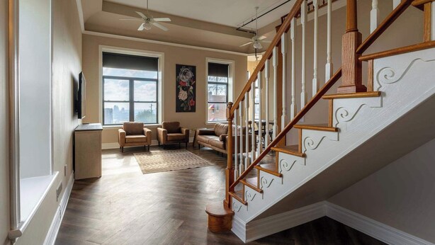 A spacious living area with a wooden staircase, large windows, and a ceiling fan.