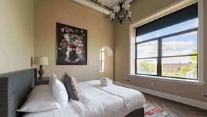 A bedroom with a large window, a bed with white linens, a painting on the wall, and a chandelier.