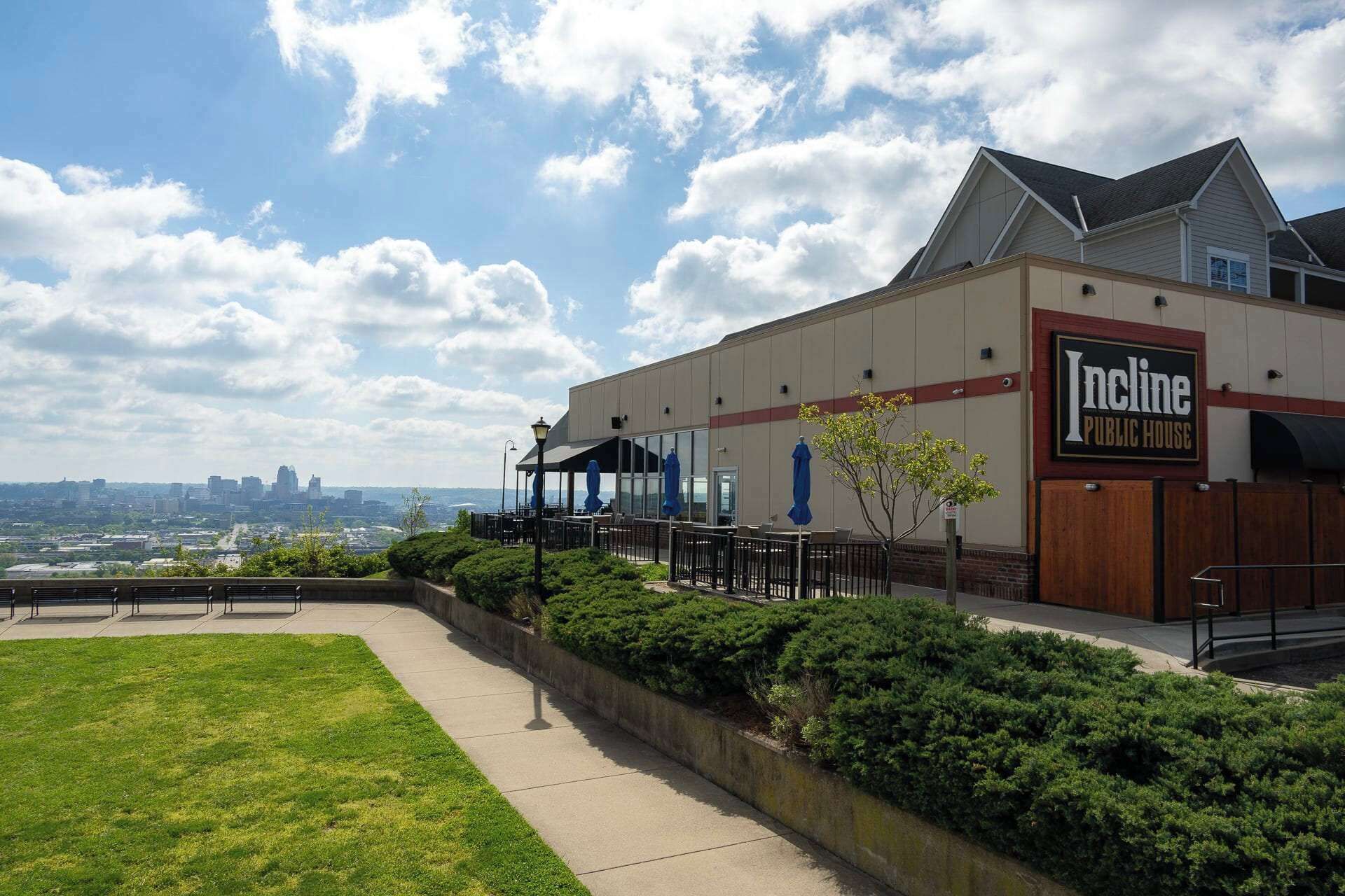 A public house with a covered outdoor seating area, a view of the city skyline, and a well-maintained lawn.
