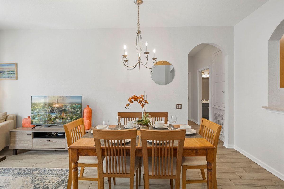 Un comedor con una mesa de madera dispuesta para cuatro, una lámpara de araña y un televisor sobre un mueble.