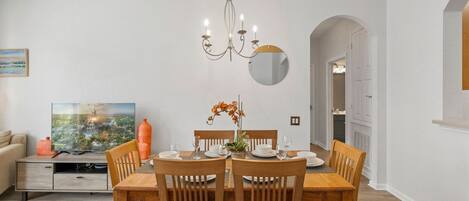 Un comedor con una mesa de madera dispuesta para cuatro, una lámpara de araña y un televisor sobre un mueble.