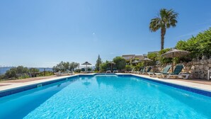 En rektangulär pool med klart, blått vatten, omgiven av solstolar och parasoller, med en stenmur och palmer i bakgrunden.