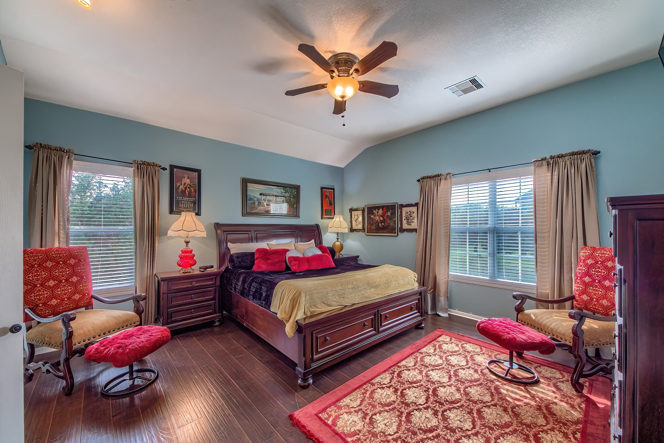 A bedroom with a bed, two chairs, a ceiling fan, and a window with curtains.