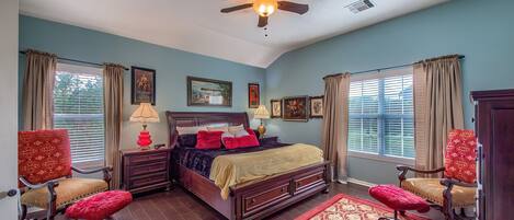 A bedroom with a bed, two chairs, a ceiling fan, and a window with curtains.