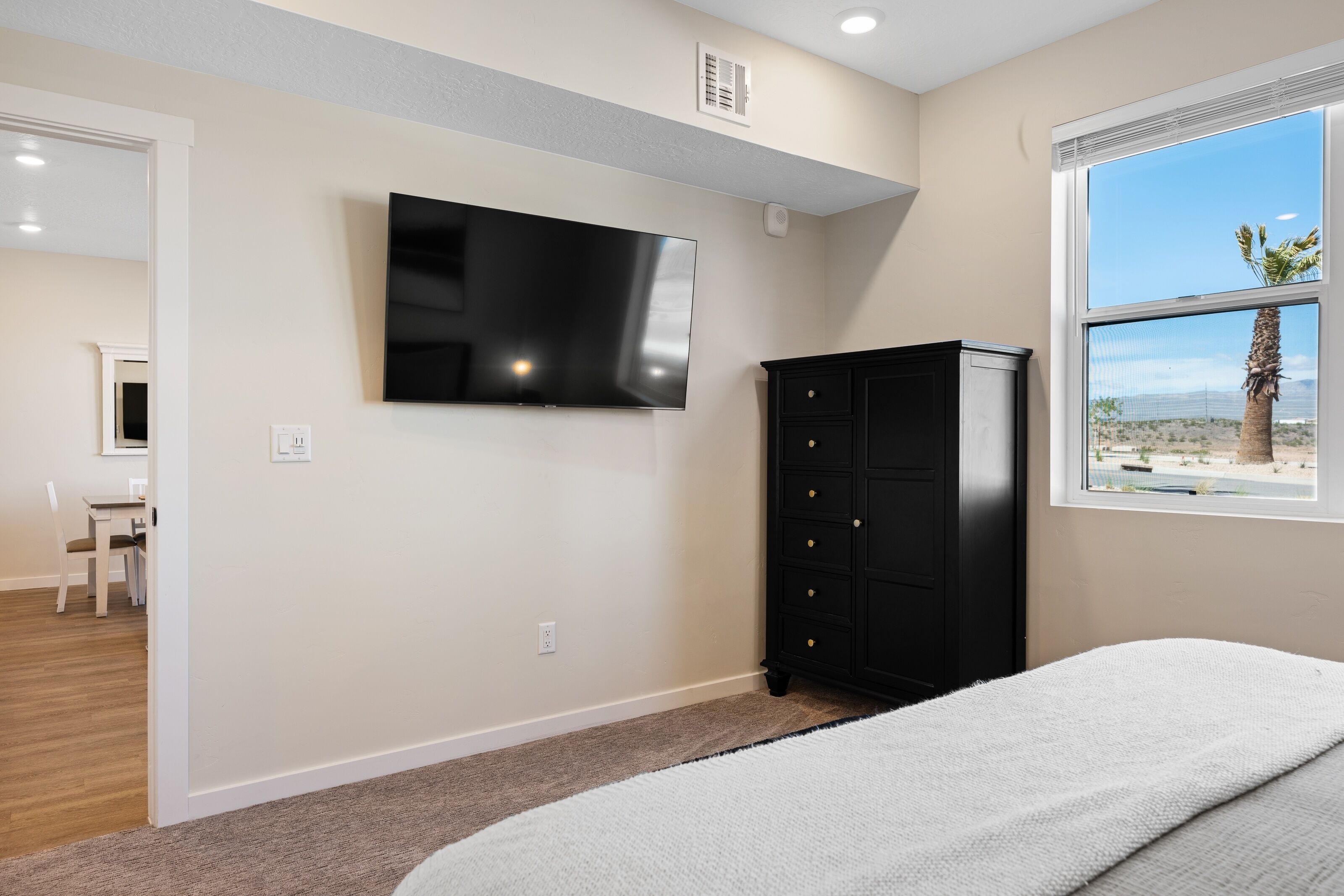 Ein Schlafzimmer mit Bett, einem an der Wand befestigten Fernseher, einem Schminktisch und einem Fenster mit Blick auf eine Wüstenlandschaft.