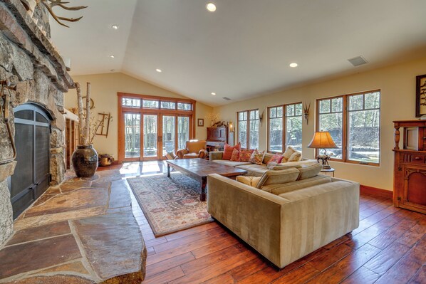 A spacious living room with a stone fireplace, a large sofa, a dining table, and large windows offering a view of the outdoors.