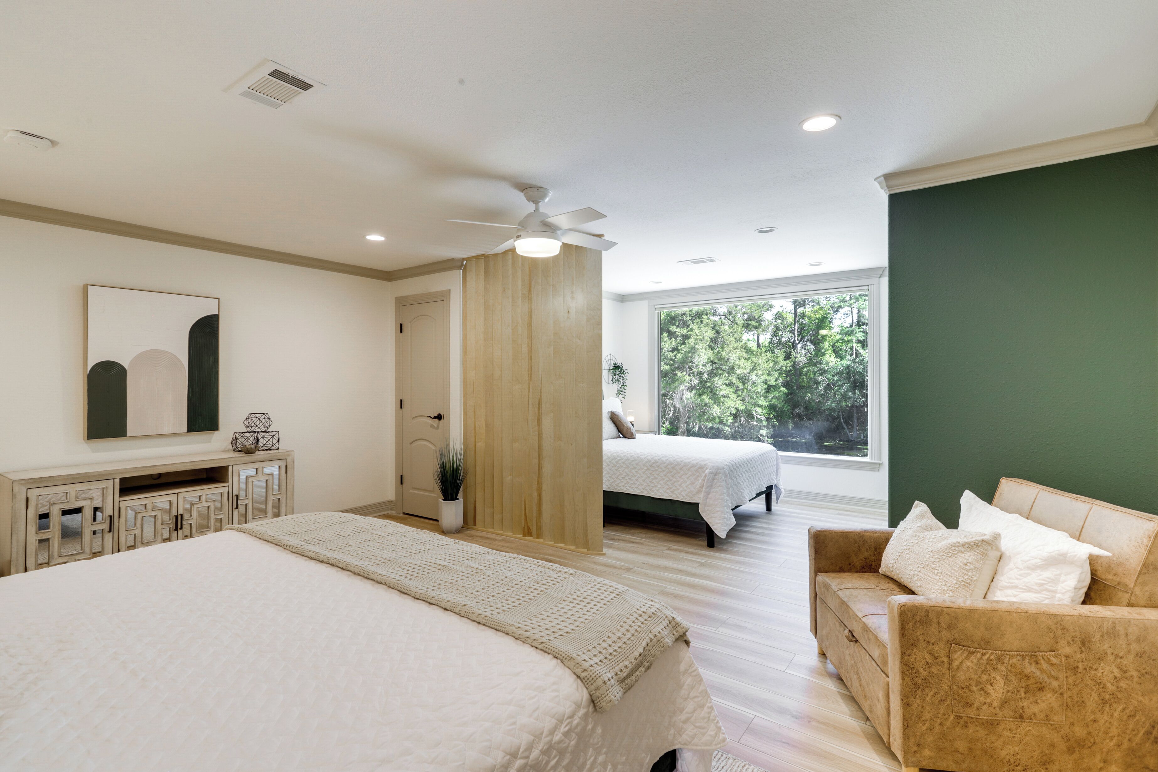 Une chambre avec un grand lit, un plus petit lit, un canapé, un ventilateur de plafond et une fenêtre donnant sur des arbres.