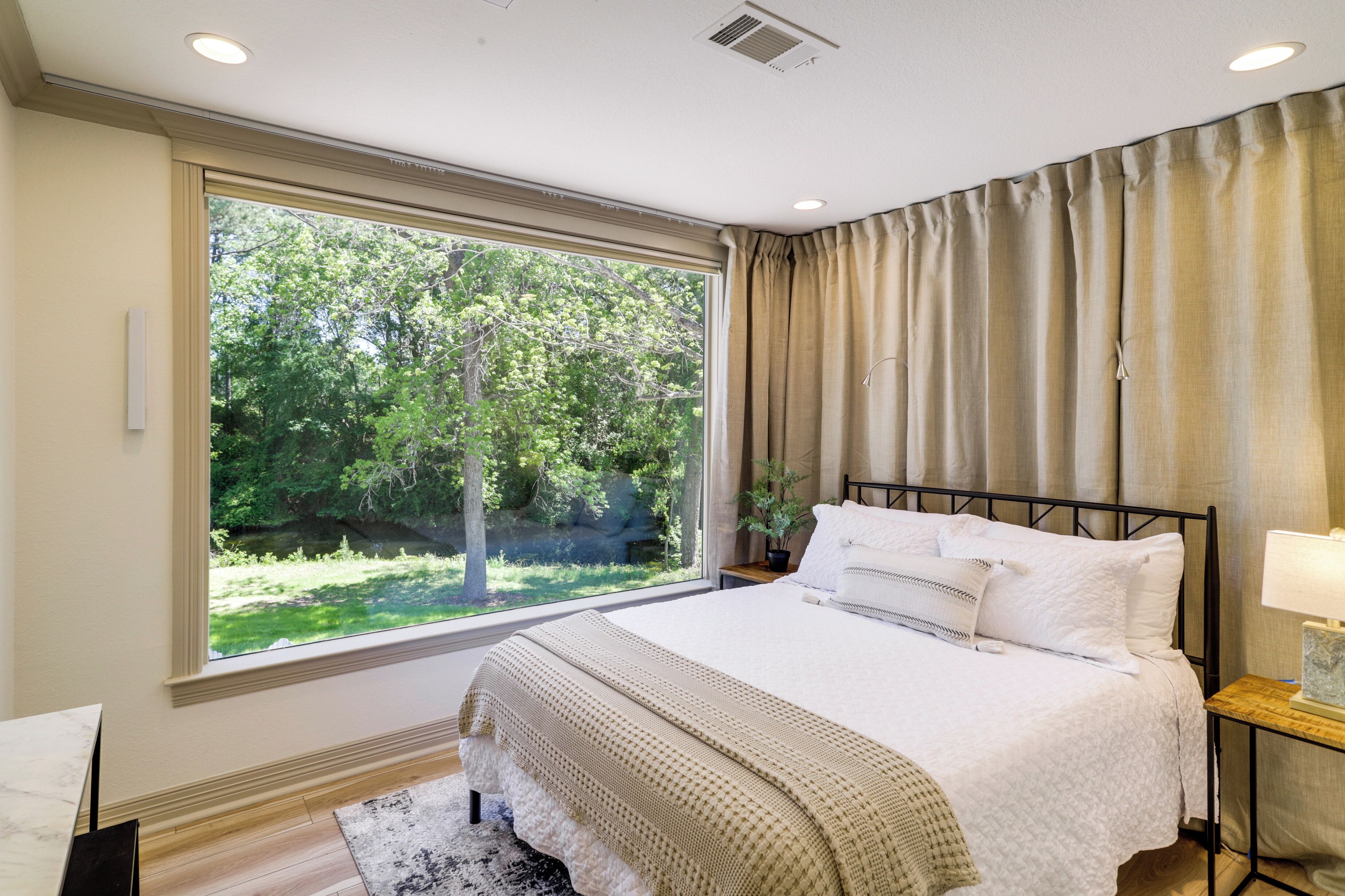 A bedroom with a large window, a bed with white bedding, a bedside table with a lamp, and a view of greenery outside.