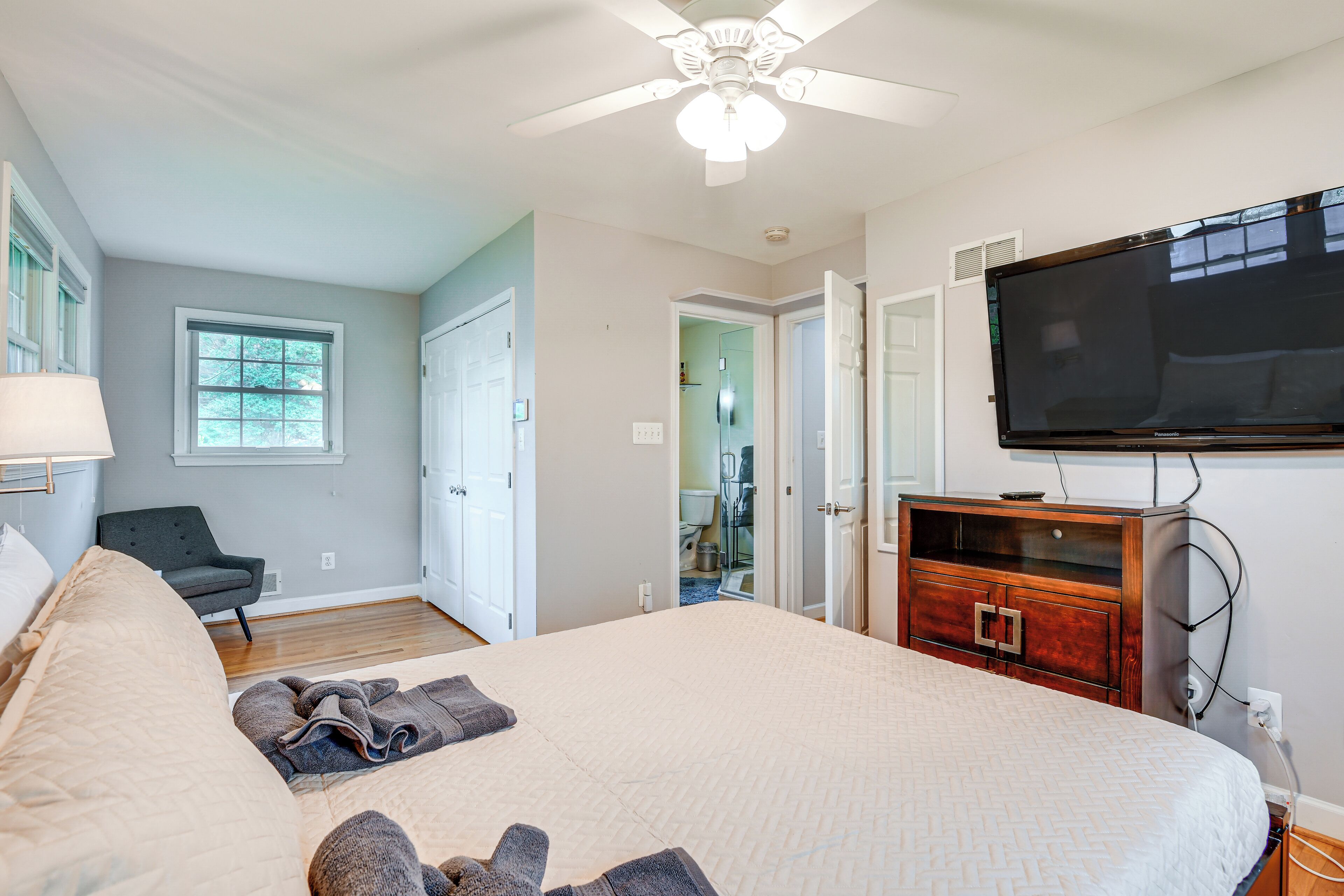 A bedroom with a large bed, a ceiling fan, a TV mounted on the wall, and a wooden dresser.