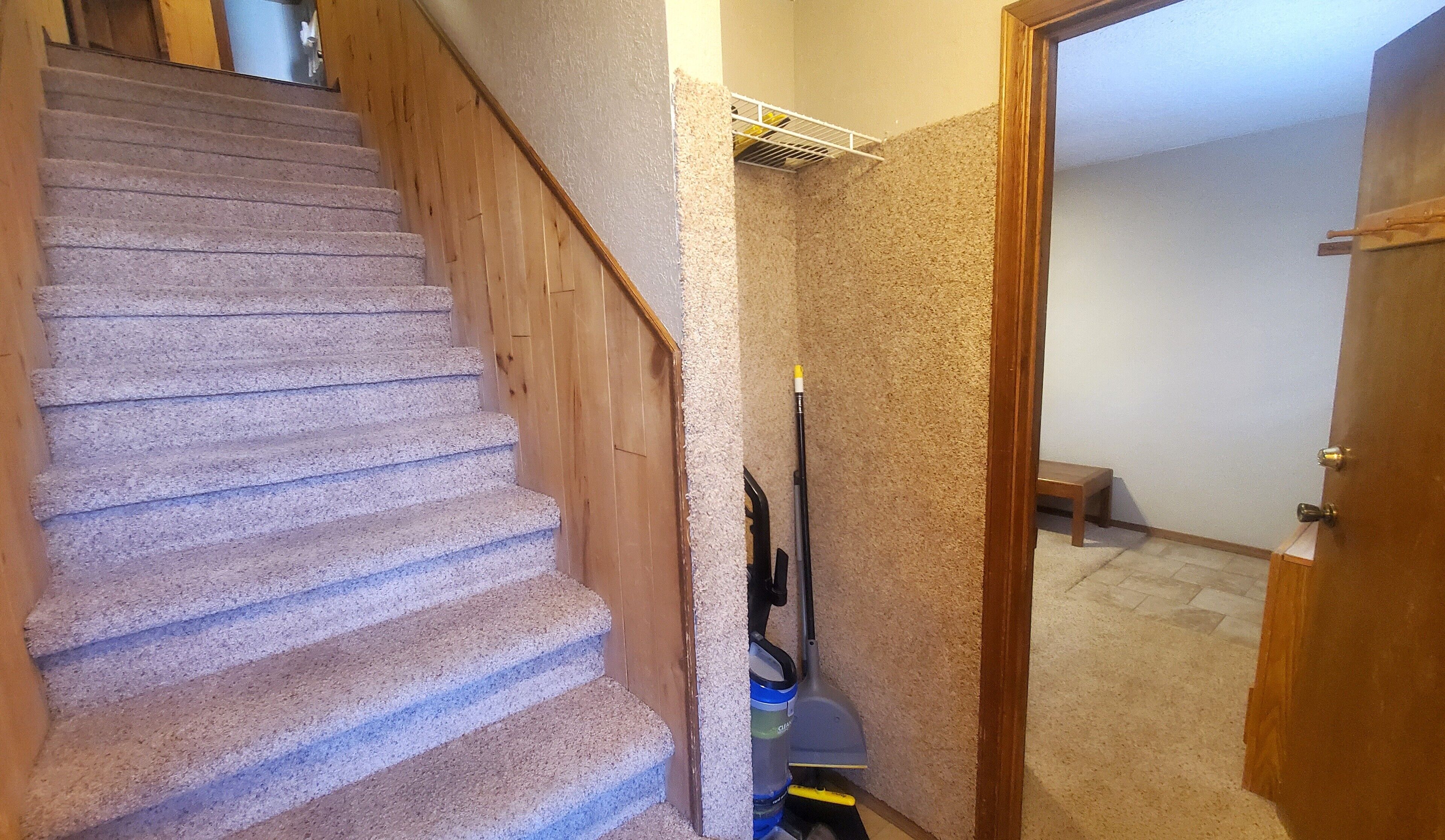 Un escalier recouvert de moquette, avec des rampes en bois, un aspirateur et une porte donnant sur une pièce dont le sol est carrelé.