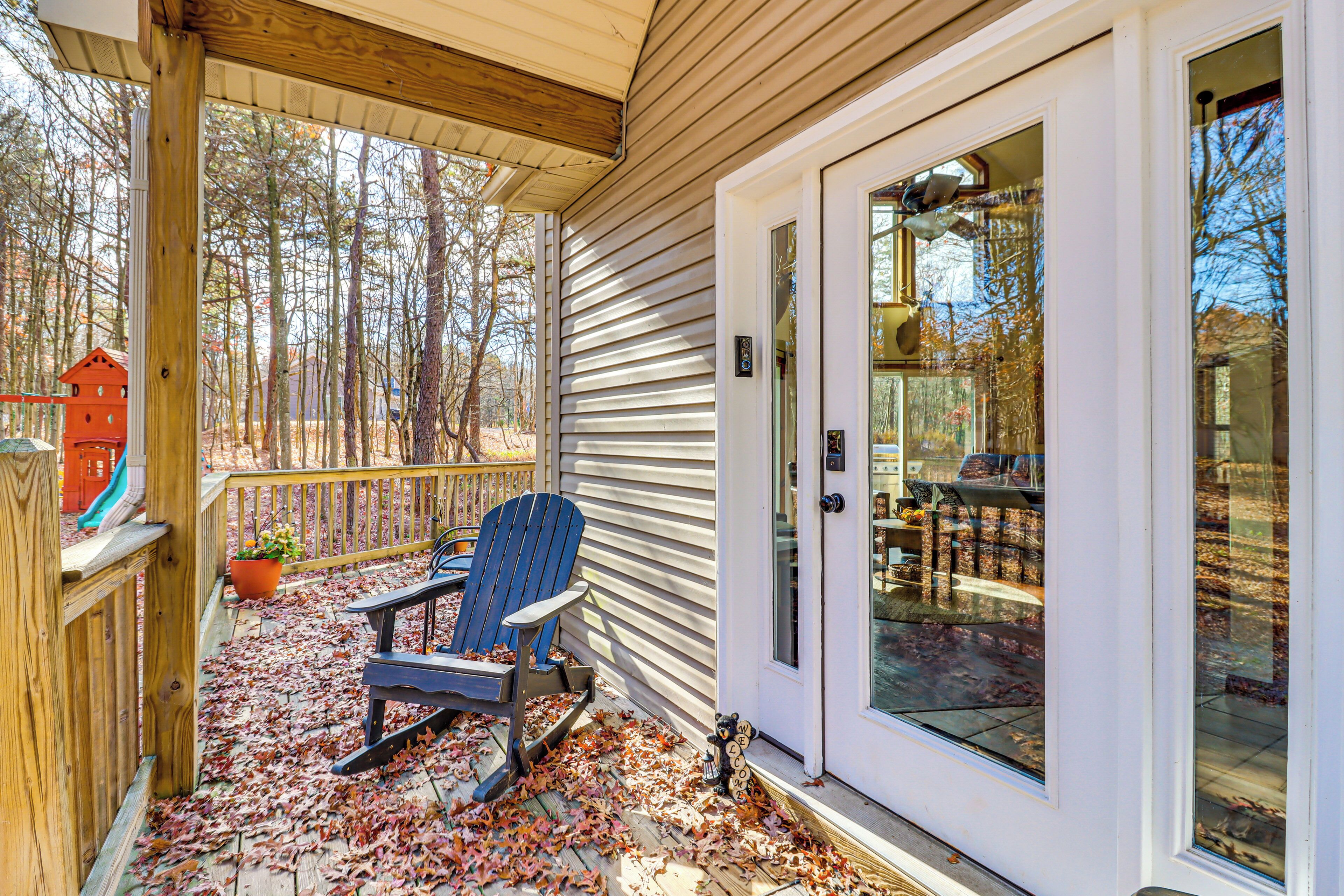 A covered porch with a blue Adirondack chair, a wooden deck, and a glass door leading to a dining area.