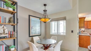 A dining area with a round glass table and four white chairs, a hanging chandelier, and a bookshelf with books and plants.