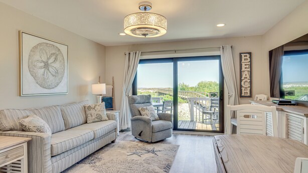 A spacious living room with a sofa, armchair, and a dining table. A large window offers a view of the outdoors.