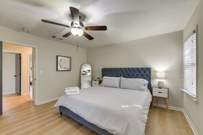 A bedroom with a large bed, a ceiling fan, a bedside table with a lamp, and a window with blinds.