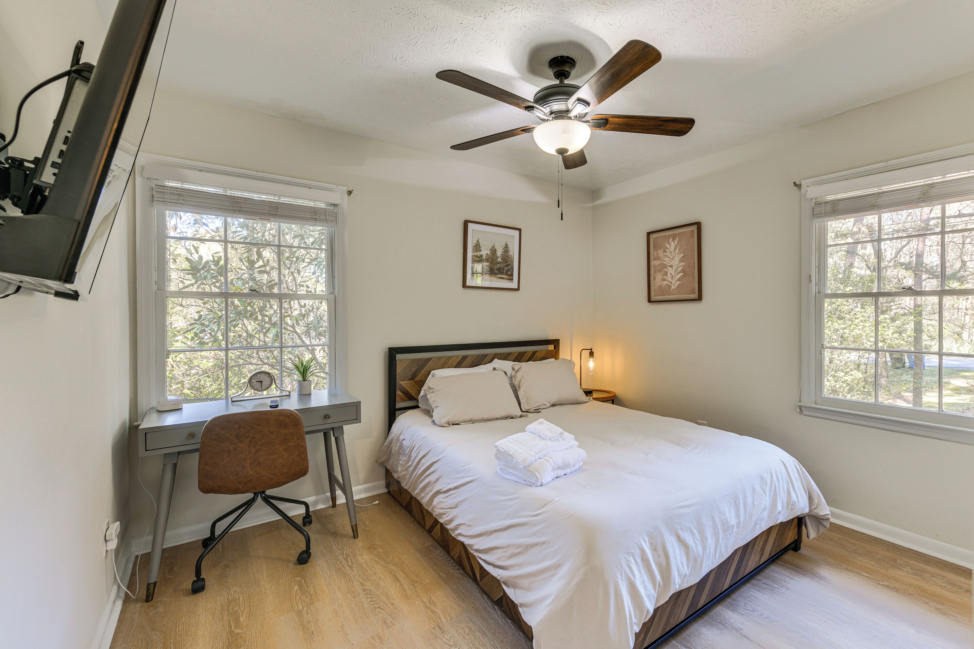 Quarto com cama, escrivaninha, cadeira, ventilador de teto e duas janelas.